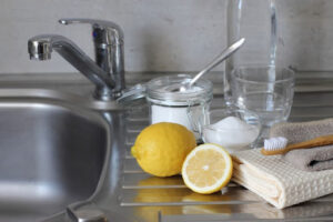 Jar of baking soda with spoon, 1 and half lemon, glass of water, towels, and toothbrush resting next to kitchen sink as homeowner prepare to unclog drain.